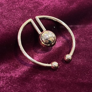 May include: A gold-toned metal ring with a circular design and a small sphere in the centre. The ring has two small spheres on the ends of the curved arms.