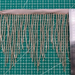 May include: A close-up of a gold beaded fringe trim with a beige backing. The fringe is approximately 1 inch (2.5 cm) long.