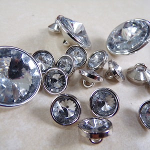 May include: A selection of clear, faceted crystal buttons with silver-coloured metal rims and shanks. The buttons vary in size and shape, with round and oval designs. The crystals sparkle, reflecting light.