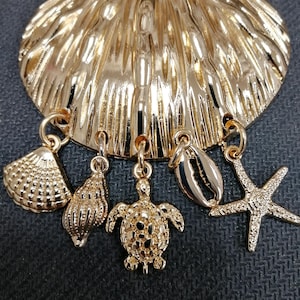 May include: A gold-coloured seashell pendant with five dangling charms: a seashell, a spiral shell, a turtle, a clam shell, and a starfish. The pendant has a textured surface against a dark background.