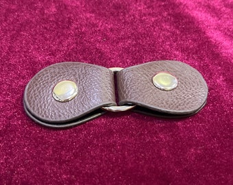 May include: A brown leather strap with two metal rings and two gold-toned metal buttons. The strap is designed to be used as a closure for a bag or other item.