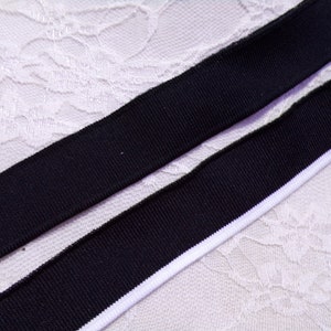 Puede incluir: Dos bandas elásticas negras con ribete blanco, sobre una superficie de encaje blanco.