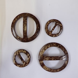 May include: Four brown coconut shell belt buckles of varying sizes. The buckles are round with a centre opening and a horizontal bar across the opening.