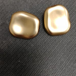 Puede incluir: Dos botones dorados de forma irregular. Los botones tienen una superficie lisa y reflectante y una forma ligeramente redondeada. Se encuentran sobre un fondo gris oscuro texturizado.