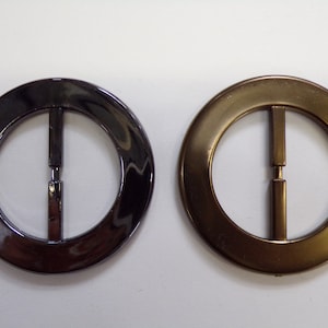 Könnte beinhalten: Zwei runde Metall-Gürtelschnallen, eine schwarze und eine goldene. Beide Schnallen haben eine Mittelstange.
