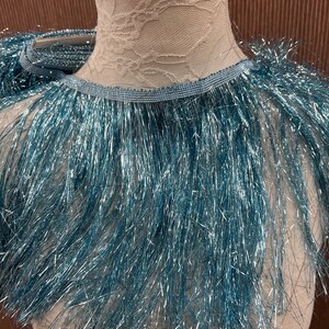 May include: A turquoise blue fringe trim with a metallic sheen. The fringe is approximately 1 inch long and is attached to a thin, blue ribbon.