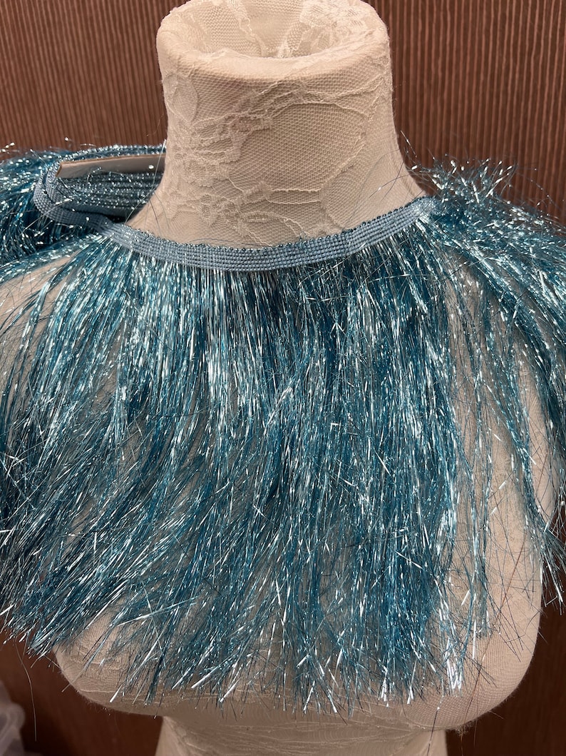 May include: A turquoise blue fringe trim with a metallic sheen. The fringe is approximately 1 inch long and is attached to a thin, blue ribbon.