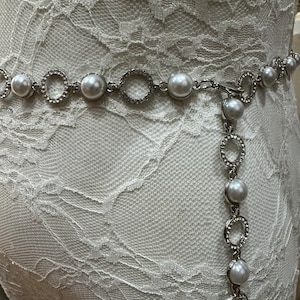 May include: A silver chain belt with faux pearls and rhinestone accents. The belt is on a white lace background.