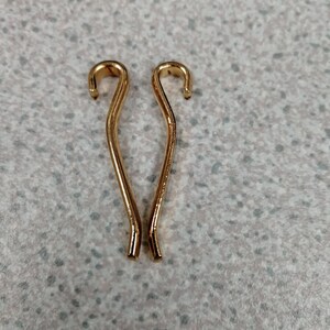 May include: Two gold-coloured hair pins with a curved design. Each pin has a hook at the top and a straight, slender body. The pins are positioned vertically against a speckled, light grey background.