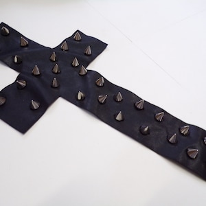 Puede incluir: Una cruz de cuero negro con tachuelas piramidales plateadas. La cruz mide aproximadamente 30 cm de largo y 15 cm de ancho.