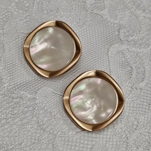 May include: A pair of gold-toned earrings with a white iridescent shell design. The earrings are round and have a slightly irregular shape.