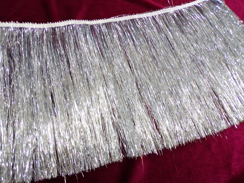May include: Silver metallic fringe trim with a white backing. The fringe is about 1 inch long.