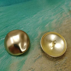 May include: Two round, gold-coloured buttons with a smooth, matte finish. One button is positioned to show its convex, domed front surface, while the other is flipped to reveal its concave underside featuring a central metal shank for attachment.