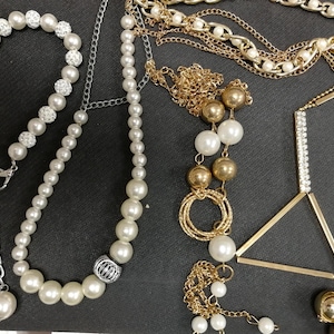 May include: A selection of necklaces and a bracelet. The jewellery features faux pearls, gold chains, and rhinestone accents. The pieces are displayed on a dark grey surface. The bracelet has a silver chain.