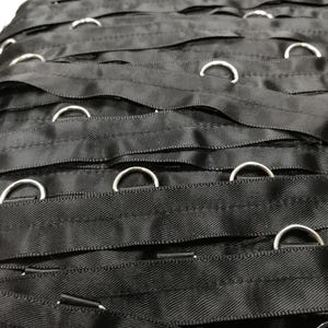 May include: A collection of black fabric ribbons arranged in parallel rows, each featuring evenly spaced silver-toned metal D-rings attached along the length of the material. The ribbons have a smooth, slightly reflective texture and are stacked horizontally.
