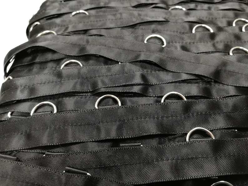 May include: A collection of black fabric ribbons arranged in parallel rows, each featuring evenly spaced silver-toned metal D-rings attached along the length of the material. The ribbons have a smooth, slightly reflective texture and are stacked horizontally.