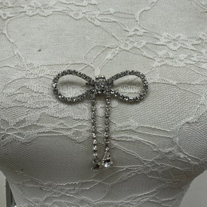 May include: A silver rhinestone bow brooch with two dangling strands of rhinestones.