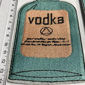 May include: A teal blue embroidered patch of a vodka bottle with a brown label that says "vodka" and a triangle symbol. The patch is about 4 inches tall.
