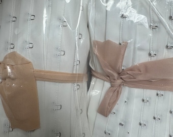 May include: Two white corset panels with metal hook-and-eye closures, wrapped in clear plastic. Each panel is tied with a beige ribbon. The corsets are likely new and unused.