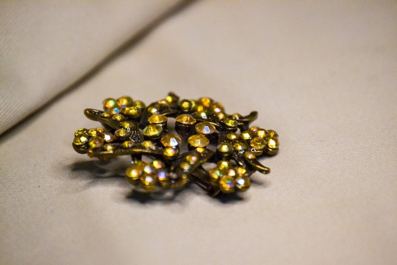 May include: A gold-toned brooch with a floral design, featuring a cluster of iridescent yellow rhinestones.