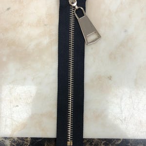 Op de afbeelding: Een zwarte rits met een zilveren trekkoord en een zilveren schuif. De rits is gesloten en het trekkoord bevindt zich bovenaan.