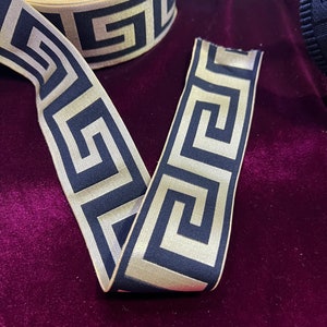 Puede incluir: Cinta decorativa con un patr&oacute;n de llave griega repetido en azul marino y dorado. La cinta se muestra sobre una superficie de terciopelo morado oscuro. La cinta est&aacute; desenrollada, mostrando el patr&oacute;n.