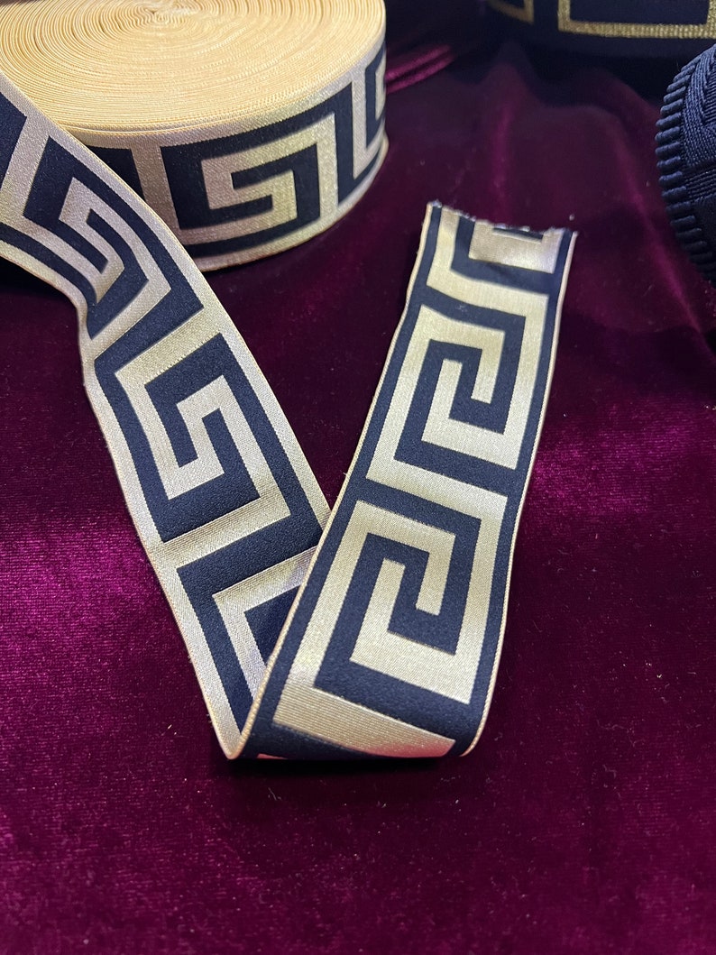 Puede incluir: Cinta decorativa con un patr&oacute;n de llave griega repetido en azul marino y dorado. La cinta se muestra sobre una superficie de terciopelo morado oscuro. La cinta est&aacute; desenrollada, mostrando el patr&oacute;n.