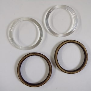 Peut inclure: Quatre anneaux circulaires sont présentés sur un fond blanc. Deux sont transparents et clairs, possiblement en plastique. Les deux autres sont bruns, avec une texture semblable au bois. Ces anneaux pourraient servir pour l'artisanat ou la bijouterie.