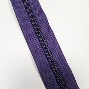 May include: A close-up of a purple zipper tape with a black zipper pull. The zipper tape is made of a woven fabric and has a subtle texture.