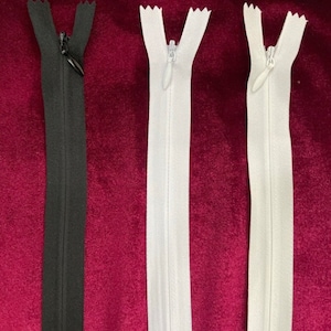 May include: Three zipper closures, one black, two white, laid out on a burgundy fabric background.