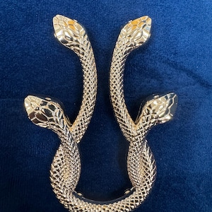 Puede incluir: Un par de asas en forma de serpiente de color dorado con una superficie texturizada. Las asas están entrelazadas y tienen un diseño decorativo.