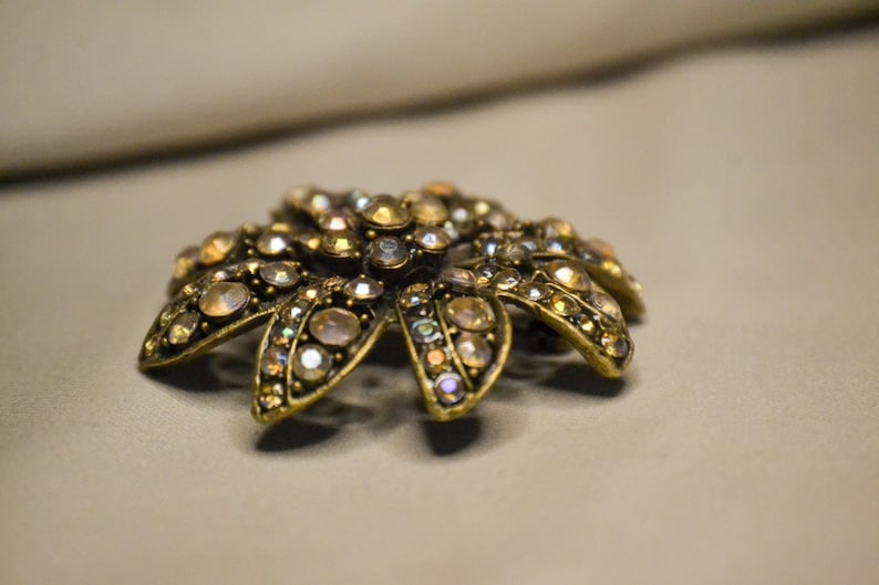 Puede incluir: Un broche de tono dorado con un dise&ntilde;o de flor, con diamantes de imitaci&oacute;n iridiscentes.
