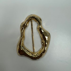 Pode incluir: Uma fivela de cinto de metal dourado com uma forma abstrata e orgânica.
