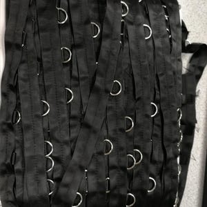 May include: A continuous roll of black satin ribbon featuring evenly spaced silver-toned metal D-rings attached along the length. The ribbon is wound in multiple vertical layers, showcasing the consistent placement of the metallic hardware.