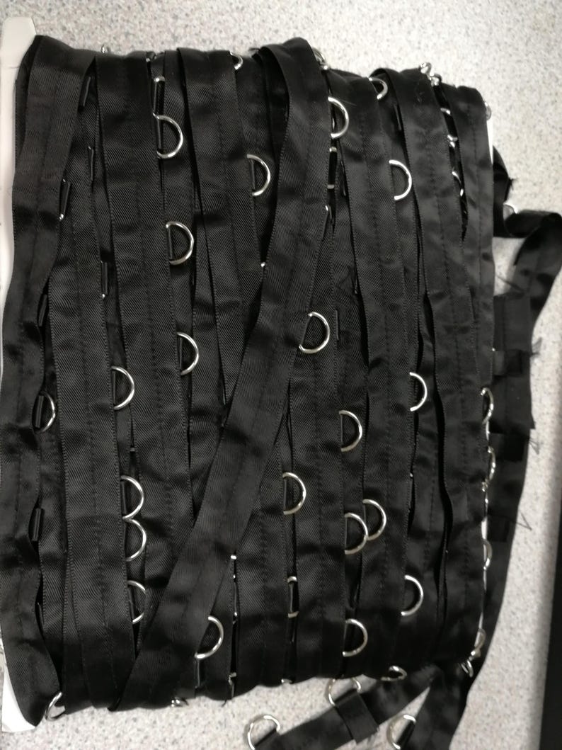 May include: A continuous roll of black satin ribbon featuring evenly spaced silver-toned metal D-rings attached along the length. The ribbon is wound in multiple vertical layers, showcasing the consistent placement of the metallic hardware.
