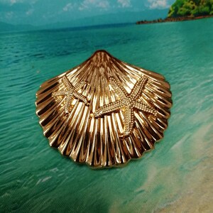 Pode incluir: Um item decorativo em forma de concha dourada com dois detalhes de estrela do mar. O item tem uma superfície texturizada e é colocado em um cenário de praia com água turquesa e céu azul.