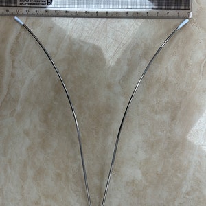 Könnte beinhalten: Eine silberne V-förmige Drahtform aus Metall, etwa 30 cm lang, mit abgerundeten Enden. Die Drahtform liegt auf einer hellbraunen Oberfläche.