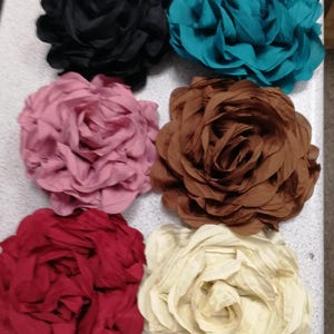 May include: Six large fabric flower embellishments in various colours: black, teal, pink, brown, red, and cream. Each flower is made of layered, ruffled fabric, resembling a rose. Suitable for crafts or fashion accessories.