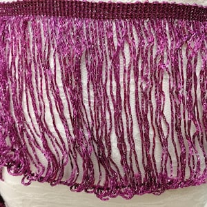 May include: Close-up of a decorative purple fringe trim. The trim has a woven top and long, flowing strands of fringe. The fringe has a metallic sheen, adding sparkle. Suitable for embellishing clothing or accessories.