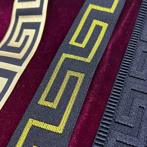 Puede incluir: Primer plano de tres cintas decorativas con patrones geom&eacute;tricos. Una cinta es negra con un dise&ntilde;o de llave griega dorada, otra es negra con un dise&ntilde;o de llave griega amarilla y la tercera es negra con un dise&ntilde;o de llave griega texturizada. Las cintas est&aacute;n sobre una superficie de terciopelo burdeos.
