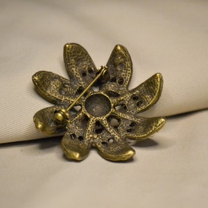 Puede incluir: Un broche de metal dorado en forma de flor con una superficie texturizada y un pasador en la parte posterior.