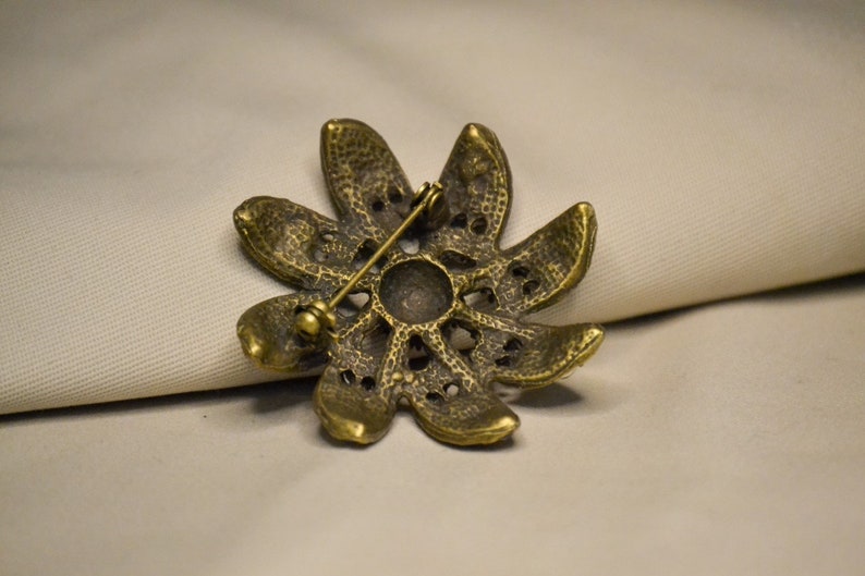 Puede incluir: Un broche de metal dorado en forma de flor con una superficie texturizada y un pasador en la parte posterior.