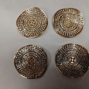May include: Four gold-colored, textured, circular buttons. Each button has a slightly irregular shape and a central design. The buttons are arranged in a square formation against a neutral background. The buttons are approximately 1.5 inches in diameter.
