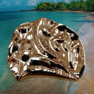 May include: A gold-coloured, irregularly shaped decorative dish with a reflective, rippled surface. The dish has a smooth, polished finish. The background shows a beach scene with blue water and sky.