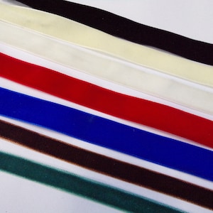 May include: Assortment of velvet ribbon in various colours, including black, white, red, blue, brown, and green. The ribbon is arranged in a diagonal pattern on a white surface.