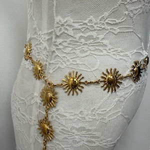Puede incluir: Cinturón dorado con abalorios en forma de sol. El cinturón está hecho de colgantes de sol unidos, con una cadena vertical de soles colgando. Se muestra sobre un fondo de encaje blanco.