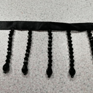 May include: A black satin ribbon trim featuring a series of dangling black beaded fringe strands. Each strand consists of multiple faceted, diamond-shaped beads that terminate in a larger, teardrop-shaped bead at the bottom edge of the trim.