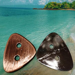 Op de afbeelding: Twee driehoekige knopen met elk twee gaten. Eén knoop is koperkleurig met een geborstelde textuur, de andere is glanzend zilverkleurig. Ze worden getoond tegen een achtergrond van een strandscène met turquoise water en een blauwe lucht.