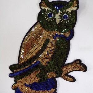 May include: A gold, green and blue sequined owl applique. The owl has large, round eyes with blue centres and white outlines. The owl's body is covered in gold sequins, with green sequins on the wings and tail. The owl's feet are blue.