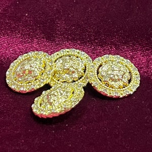 May include: Four gold-toned metal buttons with a textured, raised design. The buttons have a circular shape with a raised rim and a smaller, raised circle in the center.